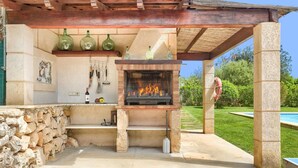 An outdoor barbecue area with a brick oven, stone walls, and a pool in the background. - Villa Joan Toni by Villa Plus (Pollença)