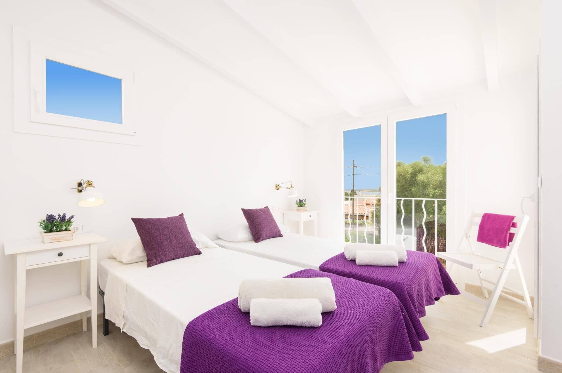 A bright, white room with two beds, each with purple bedding and white towels. There is a small table with a lamp and a plant, and a chair with a pink towel.