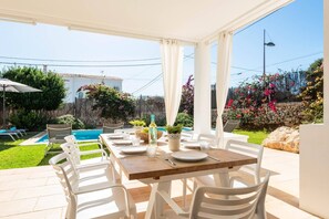 Eine Terrasse im Freien mit einem Holztisch und weißen Stühlen, ein Pool im Hintergrund und eine Mauer aus Stein mit blühenden Pflanzen.