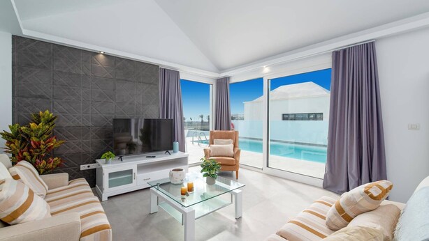 Ein modernes Wohnzimmer mit Fernseher, einem Glastisch, gestreiften Sofas und Blick auf einen Pool und ein Gebäude durch große Fenster.