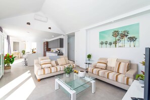 Uma sala de estar moderna com dois sofás, uma mesa de centro de vidro e uma cozinha ao fundo.
