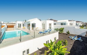 Uma área moderna de piscina no terraço, com água azul cristalina, cercada por edifícios brancos, e equipada com espreguiçadeiras e um guarda-corpo de vidro.