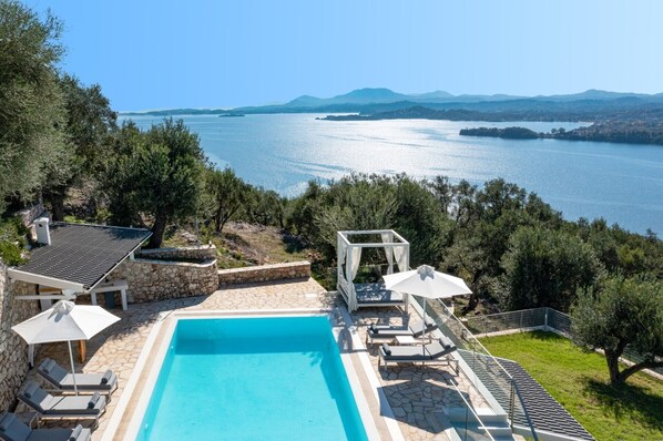 A swimming pool with lounge chairs and umbrellas, surrounded by stone walls and greenery, overlooking a large body of water and distant mountains.