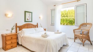A bedroom with a wooden bed, a wooden nightstand, a wicker chair, and a view of greenery through the window. - Villa Luna by Villa Plus (Cap d'Artrutx)