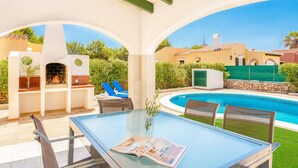 A glass-top table with a magazine, two blue lounge chairs, and a barbecue grill under a white archway. - Villa Luna by Villa Plus (Cap d'Artrutx)