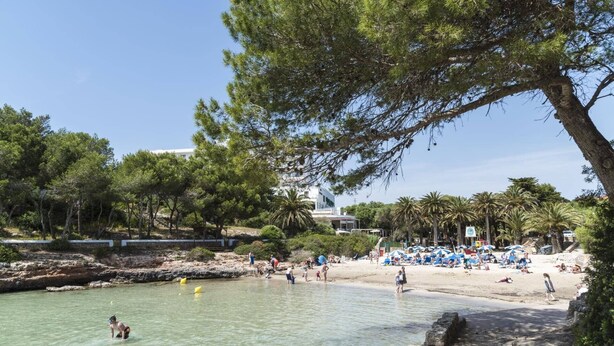 Una playa con agua cristalina, gente nadando y tomando el sol, y un edificio al fondo.