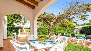 Un patio exterior con una mesa preparada para cuatro personas, rodeado de sillas blancas con cojines azules. Hay sillones de descanso junto a la piscina, y se ve una parrilla de barbacoa al fondo.