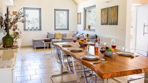 A dining area with a long wooden table set for four, a sofa with cushions, and a vase with flowers on a side table.