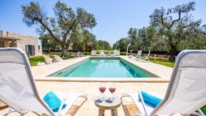A poolside view with two lounge chairs, a small table with two glasses of red wine, and a stone building in the background.