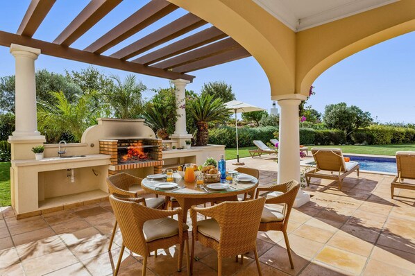 An outdoor patio with a brick grill, wicker chairs, a dining table, and a pool in the background.