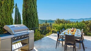 An outdoor dining area with a grill, a table set for two, and a scenic view of trees and mountains. - Villa Les Etoiles by Villa Plus (Lorgues)
