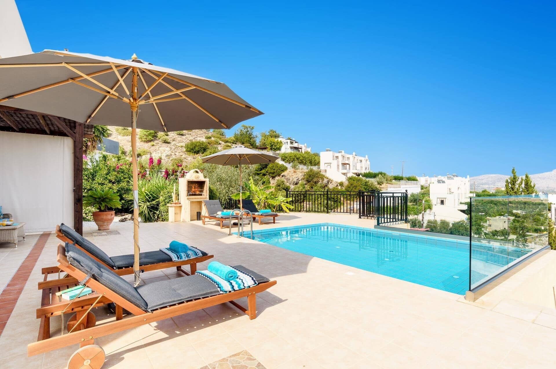 A rooftop pool area with lounge chairs and umbrellas, a barbecue grill, and a view of the surrounding hillside.