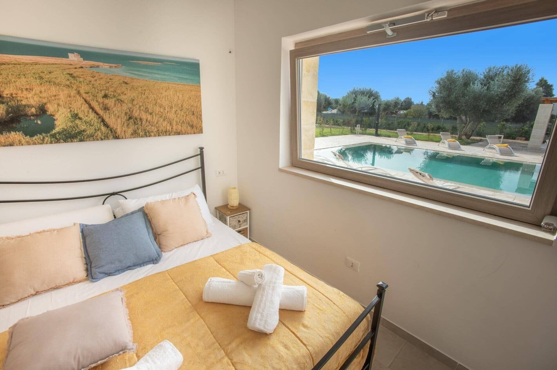 Une chambre à coucher avec un lit, des oreillers, un tableau, une fenêtre et une vue sur une piscine.