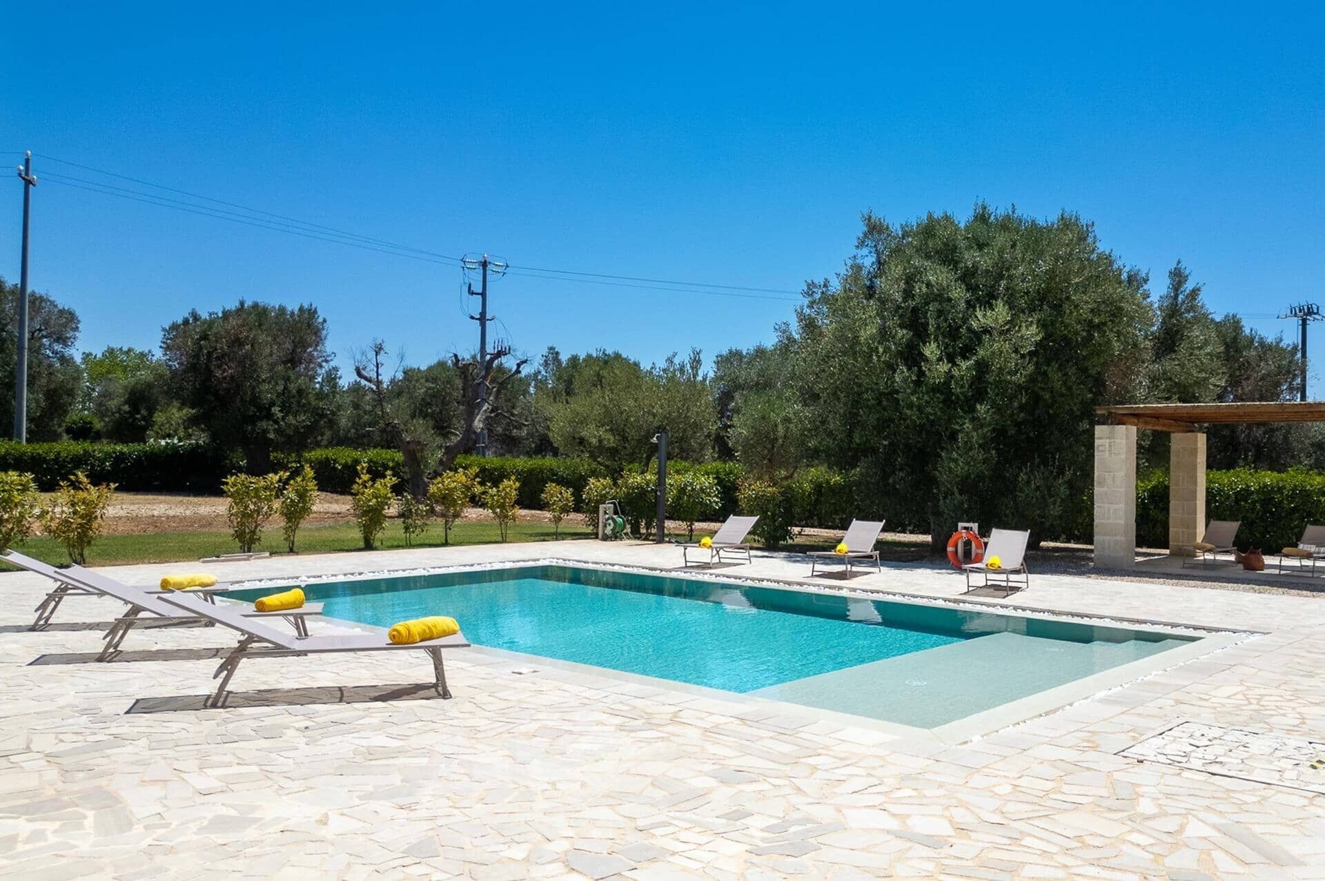 A rectangular swimming pool with a tiled deck, surrounded by lounge chairs and a pergola structure.
