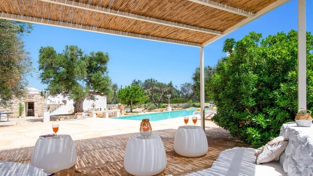 An outdoor seating area with white tables and chairs, overlooking a pool and a stone building.