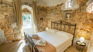 A stone-walled bedroom with a bed, a small table with a lamp, and a window with curtains.