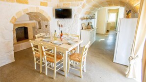 A dining area with a stone wall, a fireplace, a TV mounted on the wall, and a kitchen in the background.