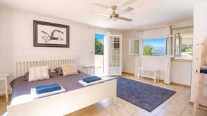 Un dormitorio con cama, ventilador de techo, cuadro enmarcado, ventana con vistas y puerta que da al exterior.