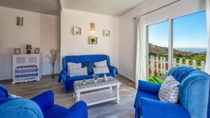 Un salon avec un canapé bleu, une table basse blanche et une vue sur un paysage côtier, visible à travers de grandes fenêtres.