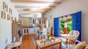 Un vaste espace de vie comprenant une table à manger, des chaises et un canapé. Il y a également un coin cuisine avec des placards et un évier. La pièce est dotée d’un sol carrelé et d’une grande fenêtre avec des portes bleues.