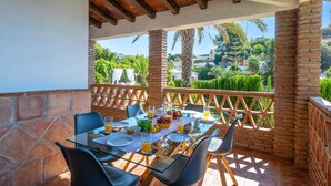 Un espace de restauration en plein air avec une table en verre, des chaises et une vue sur un jardin luxuriant et des collines.