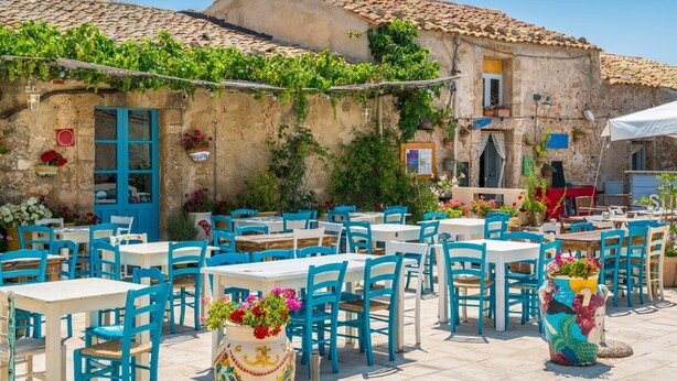 An outdoor café with tables and chairs, a stone building with a tiled roof, and potted plants. - Villa Clara Ispica by Villa Plus (Santa Maria del Focallo)