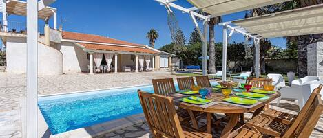 A poolside area with a covered table and chairs, a clear pool, and a building with a tiled roof in the background.