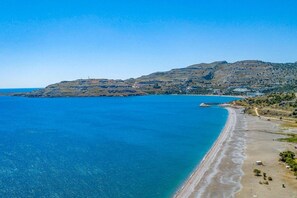 A coastal landscape with a beach, clear blue sea, and a hilly background.
