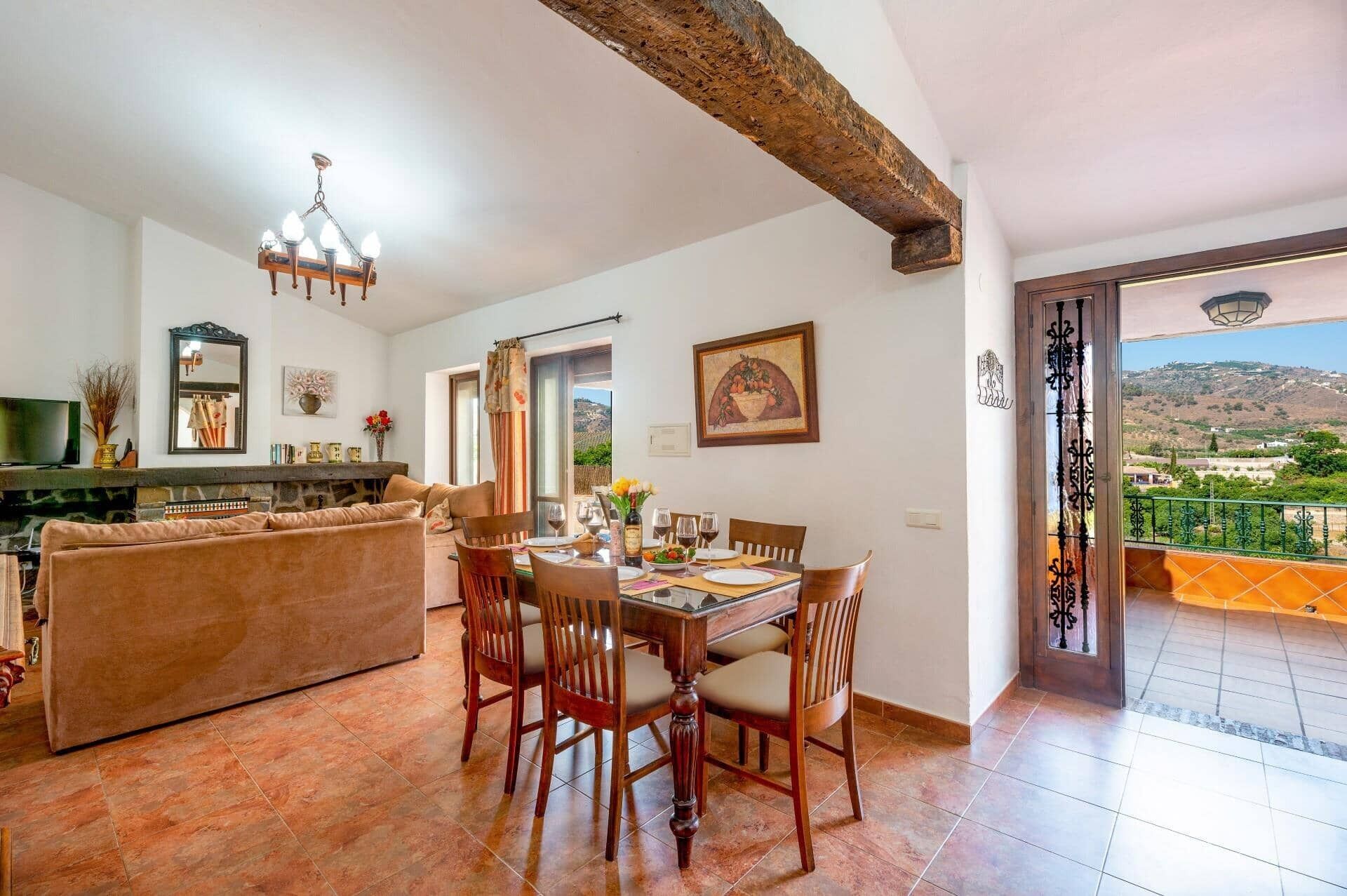 A dining area with a wooden table and chairs, a sofa, and a fireplace. There is a balcony with a view of the countryside.