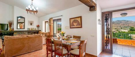 A dining area with a wooden table and chairs, a sofa, and a fireplace. There is a balcony with a view of the countryside.