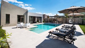 A modern outdoor pool area with lounge chairs and umbrellas, a stone wall, and a pergola with a grill. - Villa Masticha Sweet by Villa Plus (Kariotes)