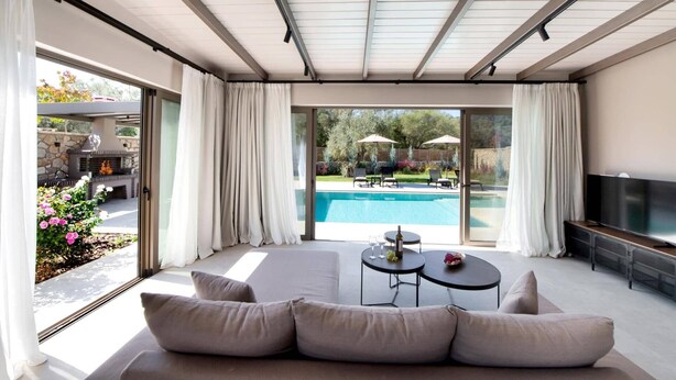 A modern living room with a large window, a pool, and outdoor furniture. - Villa Masticha Sweet by Villa Plus (Kariotes)