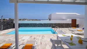 A modern outdoor pool area with a stone wall, lounge chairs, and a table set for dining, overlooking a coastal landscape. - Villa Femes by Villa Plus (Playa Blanca)