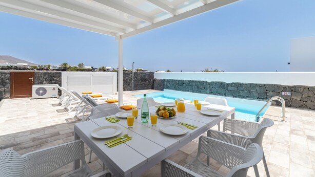 A sunlit outdoor dining area with a pool, stone walls, and a clear view of the sea. - Villa Femes by Villa Plus (Playa Blanca)