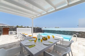 A sunlit outdoor dining area with a pool, stone walls, and a clear view of the sea. - Villa Femes by Villa Plus (Playa Blanca)