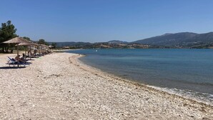 A pebble beach with beach umbrellas and lounge chairs, a clear blue sea, and distant hills. - Villa Pasteli Sweet by Villa Plus (Kariotes)
