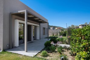 A modern building with a covered outdoor area, stone wall, and garden with various plants. - Villa Pasteli Sweet by Villa Plus (Kariotes)