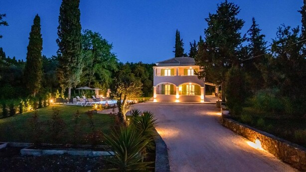 Una casa de dos pisos con un camino de entrada que lleva al acceso, rodeada de árboles y un jardín bien iluminado.