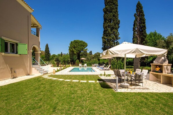 Un área de piscina con sillones de playa y sombrillas, una parrilla de barbacoa y una escalera que conduce a un edificio con contraventanas verdes.