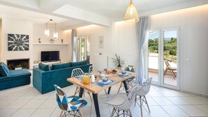 Una sala de estar moderna con un sofá, una mesa de comedor y sillas. Hay un televisor, una obra de arte en la pared y vistas al exterior a través de una puerta de cristal.