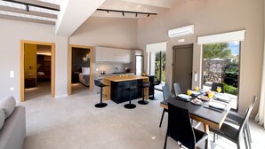 A modern kitchen with a central island, a dining area with a table set for two, and a view of the outdoors through large windows. - Villa Mandola Sweet by Villa Plus (Kariotes)