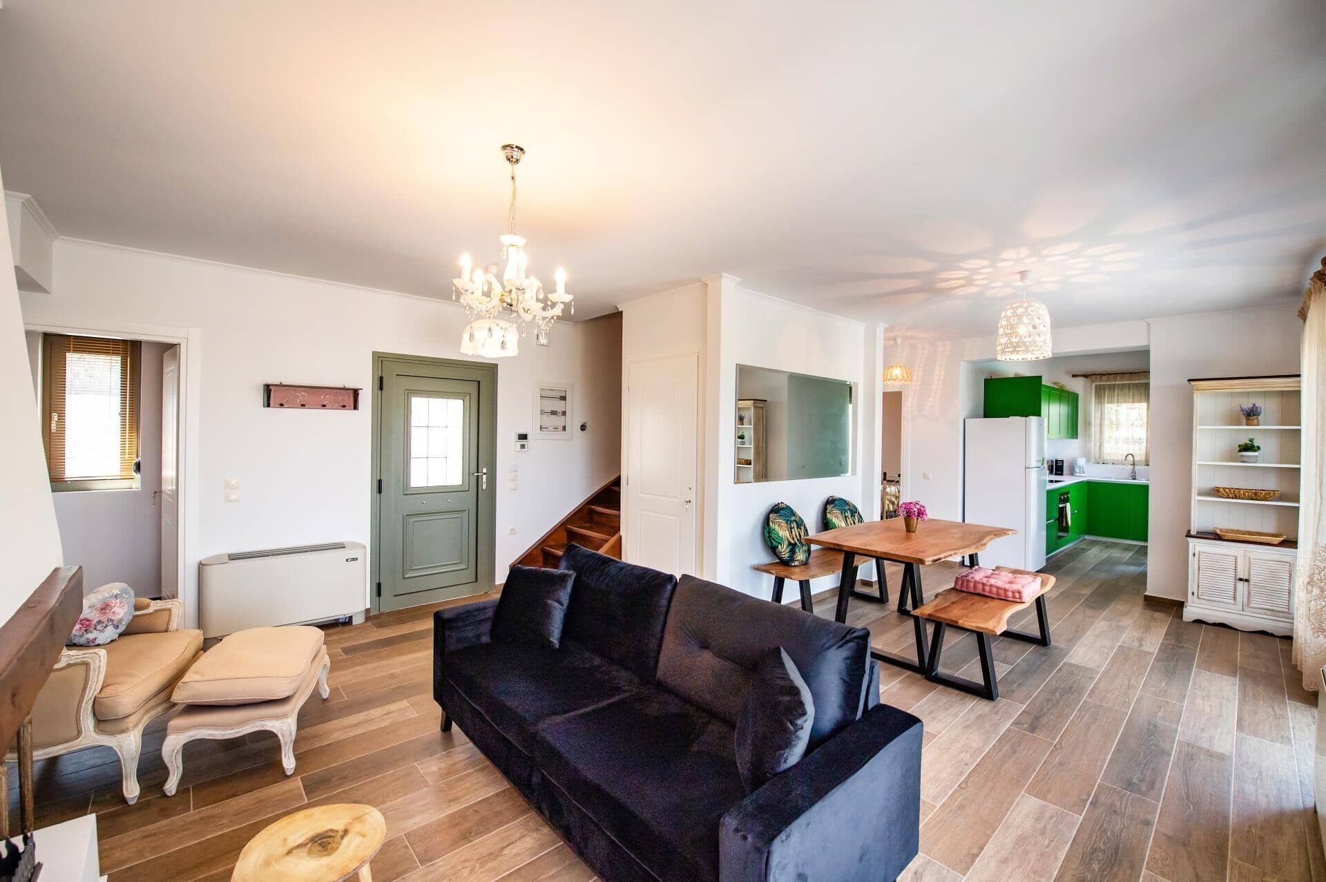 A modern living room with a dark sofa, a wooden coffee table, and a chandelier. There is a staircase leading to an upper floor, and a kitchen area with green cabinets.