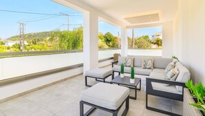 Un espace de vie extérieur moderne, doté d’un canapé, d’une table basse et d’un pouf, et offrant une vue sur la verdure et les bâtiments.