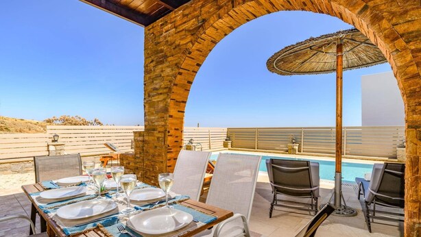 An outdoor dining area with a pool, wooden tables, and chairs, under a brick archway. - Villa Kiotari by Villa Plus (Kiotari)