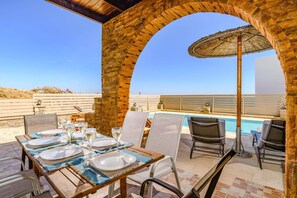 An outdoor dining area with a pool, wooden tables, and chairs, under a brick archway.