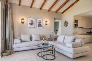 A modern living room with a sofa, a glass coffee table, and a kitchen in the background. - Villa Katoikia Dio by Villa Plus (Ionian Islands)