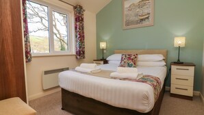 Un lit bien fait, avec du linge de lit blanc et un coussin à motifs floraux, une table de chevet avec une lampe, une fenêtre ornée de rideaux à fleurs et un tableau accroché au mur.