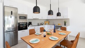 A modern kitchen with white cabinets, a stainless steel refrigerator, a microwave, and an oven. There are three pendant lights hanging above a wooden dining table set with plates, glasses, and cutlery. - Villa Malvasia by Villa Plus (Playa Blanca)