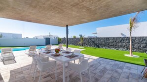 A covered outdoor dining area with a pool, white chairs, and a table set for two. - Villa Malvasia by Villa Plus (Playa Blanca)