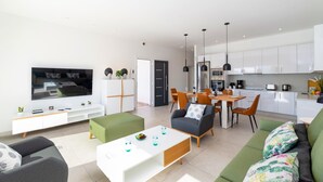 A modern living room with a flat-screen TV, a dining area with a wooden table and chairs, and a kitchen with white cabinets and appliances. - Villa Malvasia by Villa Plus (Playa Blanca)